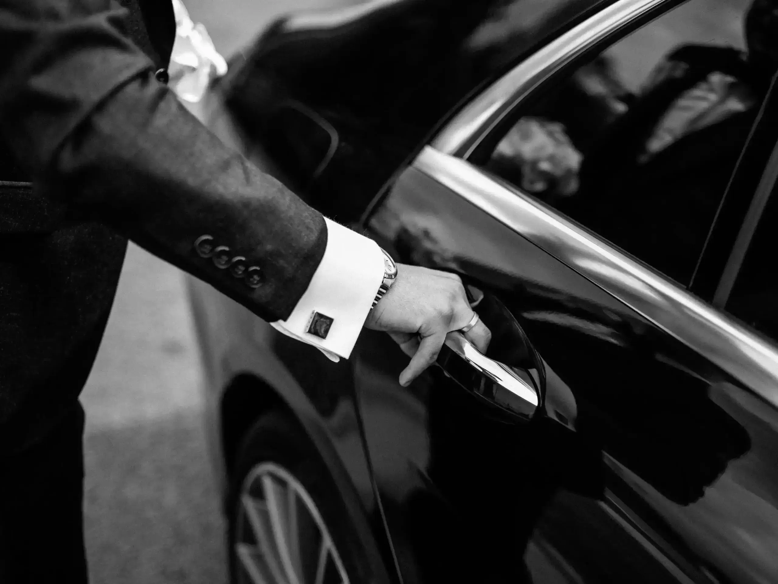 Black and white close-up of a well-dressed hand with cufflinks and a watch opening a black car door.