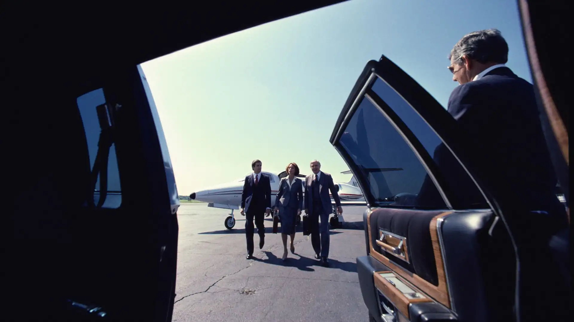 Business travelers walking toward a waiting luxury car on an airport tarmac, with a private jet in the background.