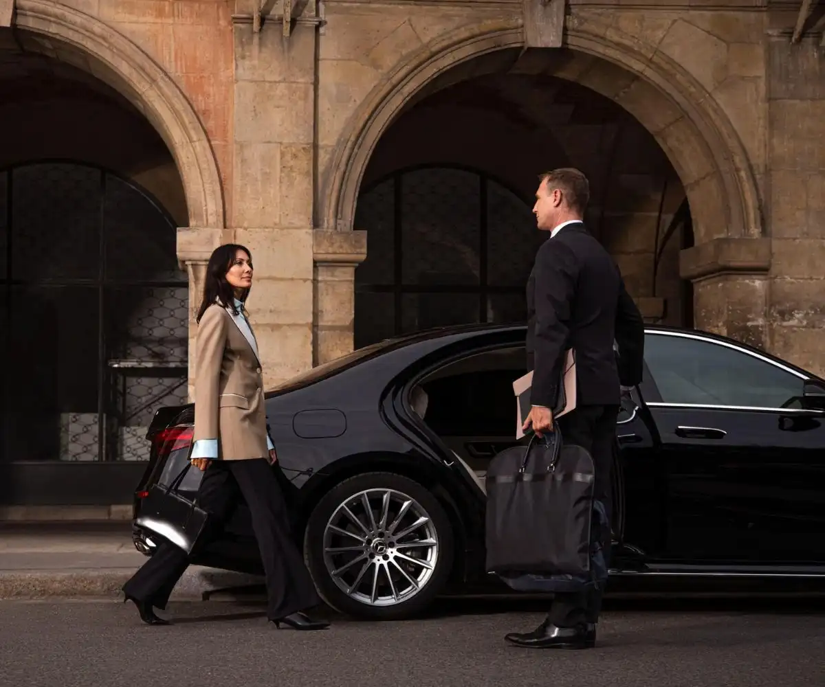 late-night-chauffeur-service Chauffeur holding a bag for a woman exiting a luxury black sedan outside a building with stone arches.