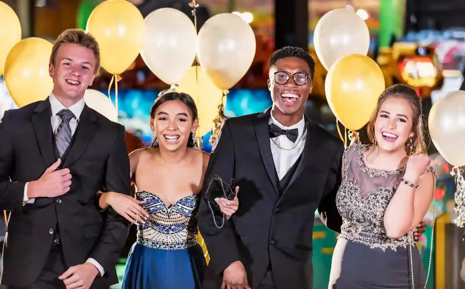 prom-night-transportation Four high school students dressed for prom smiling and linking arms in a festive setting with balloons.