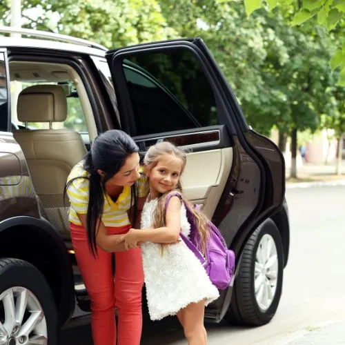 A mother helping her young daughter, who is wearing a purple backpack, step out of a luxury SUV for school