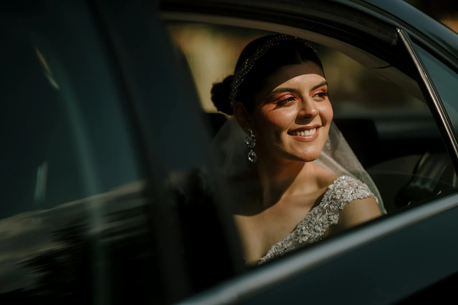 A smiling bride in a beaded white gown and jeweled headband looks out the window of a luxury black limousine on a sunny day