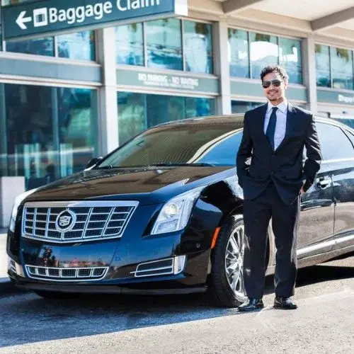 Professional chauffeur in a black suit standing next to a polished black luxury sedan at an airport baggage claim terminal