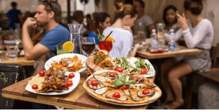 Restaurant table with pizza, pasta, salad, and drinks in a busy dining setting.